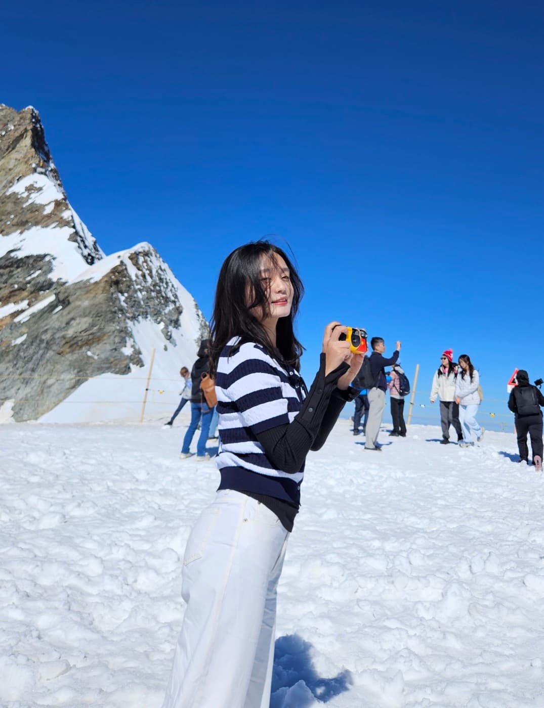 Nicole smiling and holding a small camera on a snowy mountainside, wearing a striped sweater and white pants, with a jagged snow-covered peak behind her.