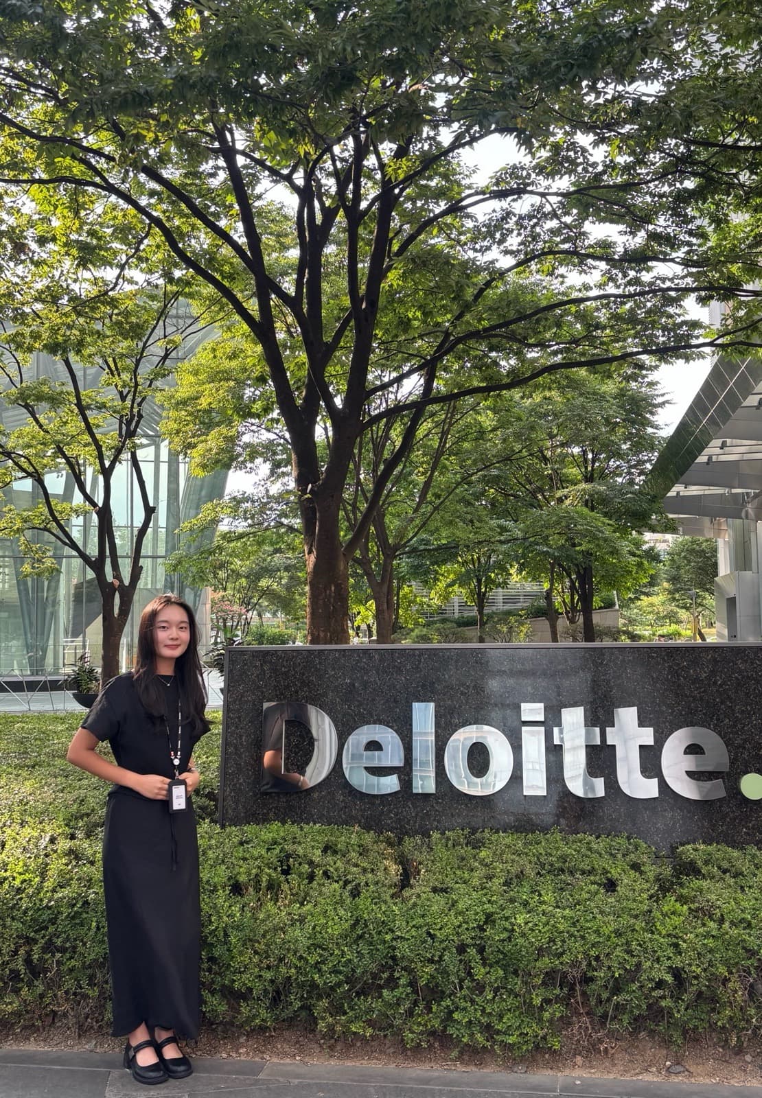 Nicole standing next to a "Deloitte." sign among trees outside the office, wearing business attire with an employee badge.