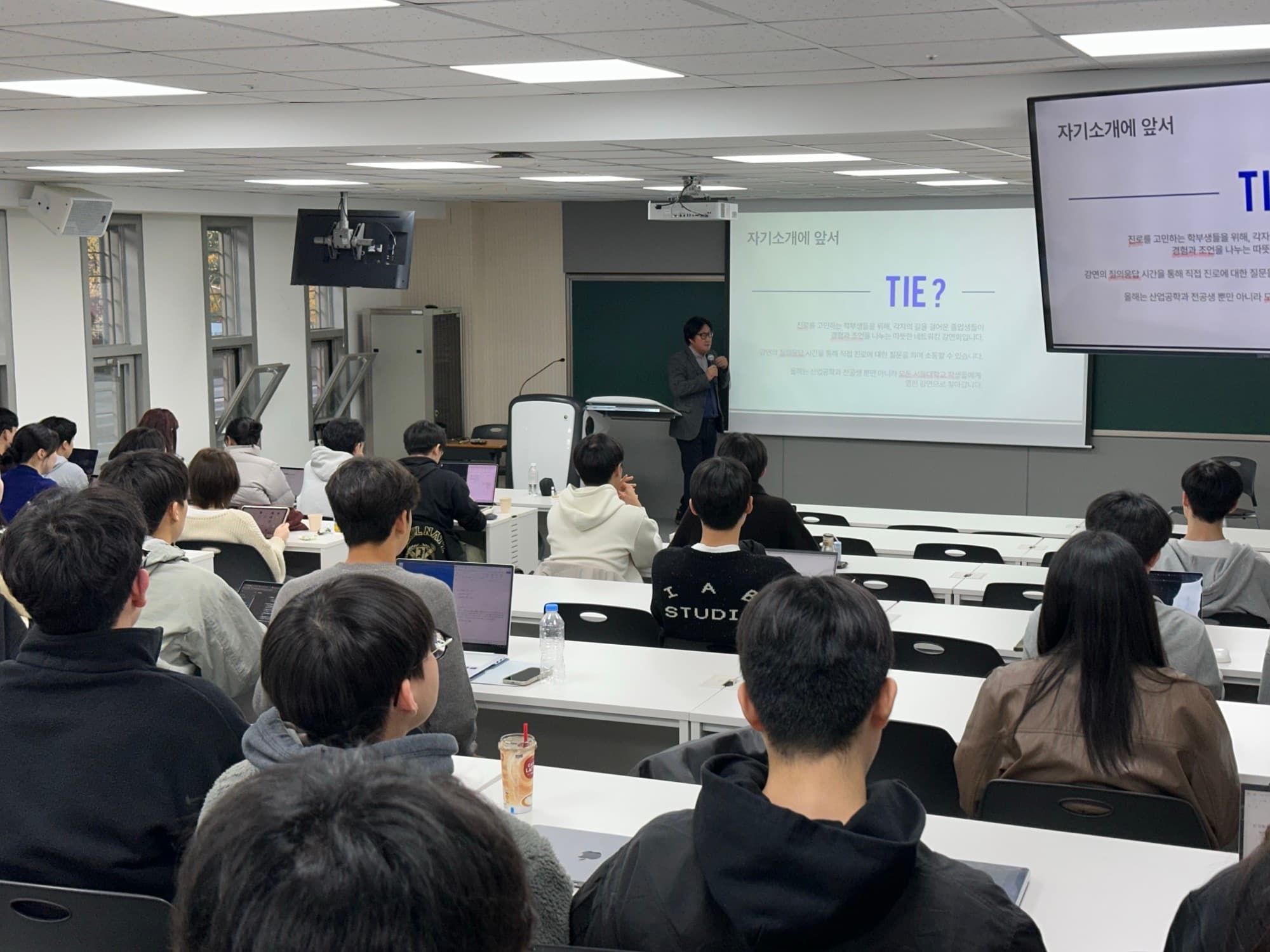 Students seated in a lecture hall listening to a speaker at the podium, in front of a screen reading "TIE" during an SNU Industrial Engineering alumni speaker event.