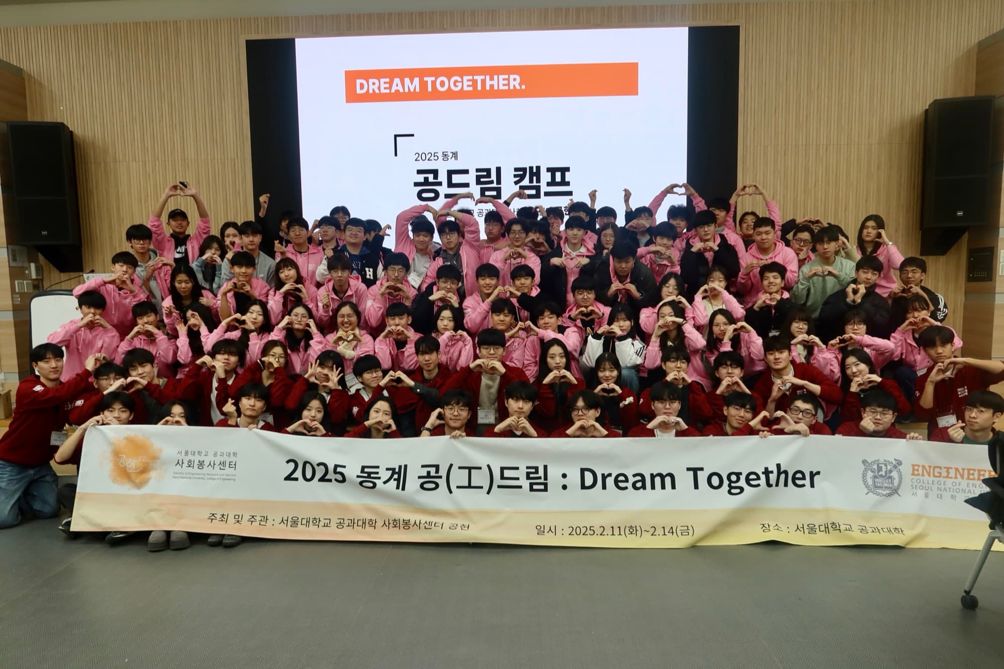 A large group of about 90 students and mentors in pink and red jackets making heart hand-shapes in an auditorium, in front of a screen reading "2025 동계 공드림 캠프: Dream Together" and a banner listing camp dates Feb 11–14, 2025.