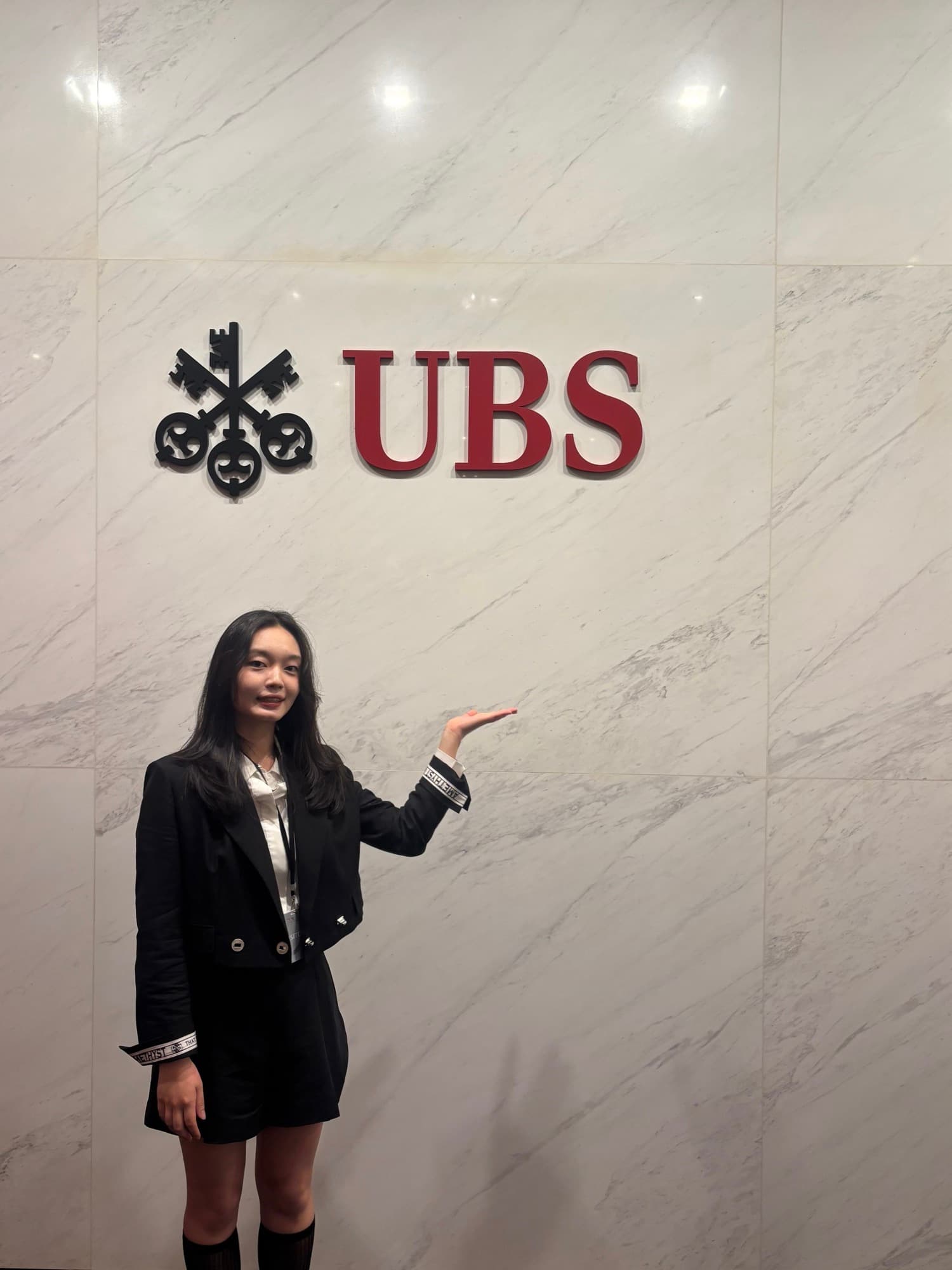 Nicole standing and gesturing toward a marble wall bearing the UBS logo and wordmark, dressed in business attire.
