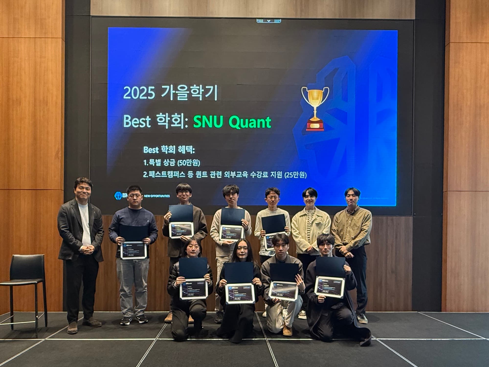 Nicole's SNU Quant team kneeling and standing on stage holding award plaques, in front of a screen reading "2025 가을학기 Best 학회: SNU Quant" (2025 Fall Semester Best Club: SNU Quant) with a trophy graphic.