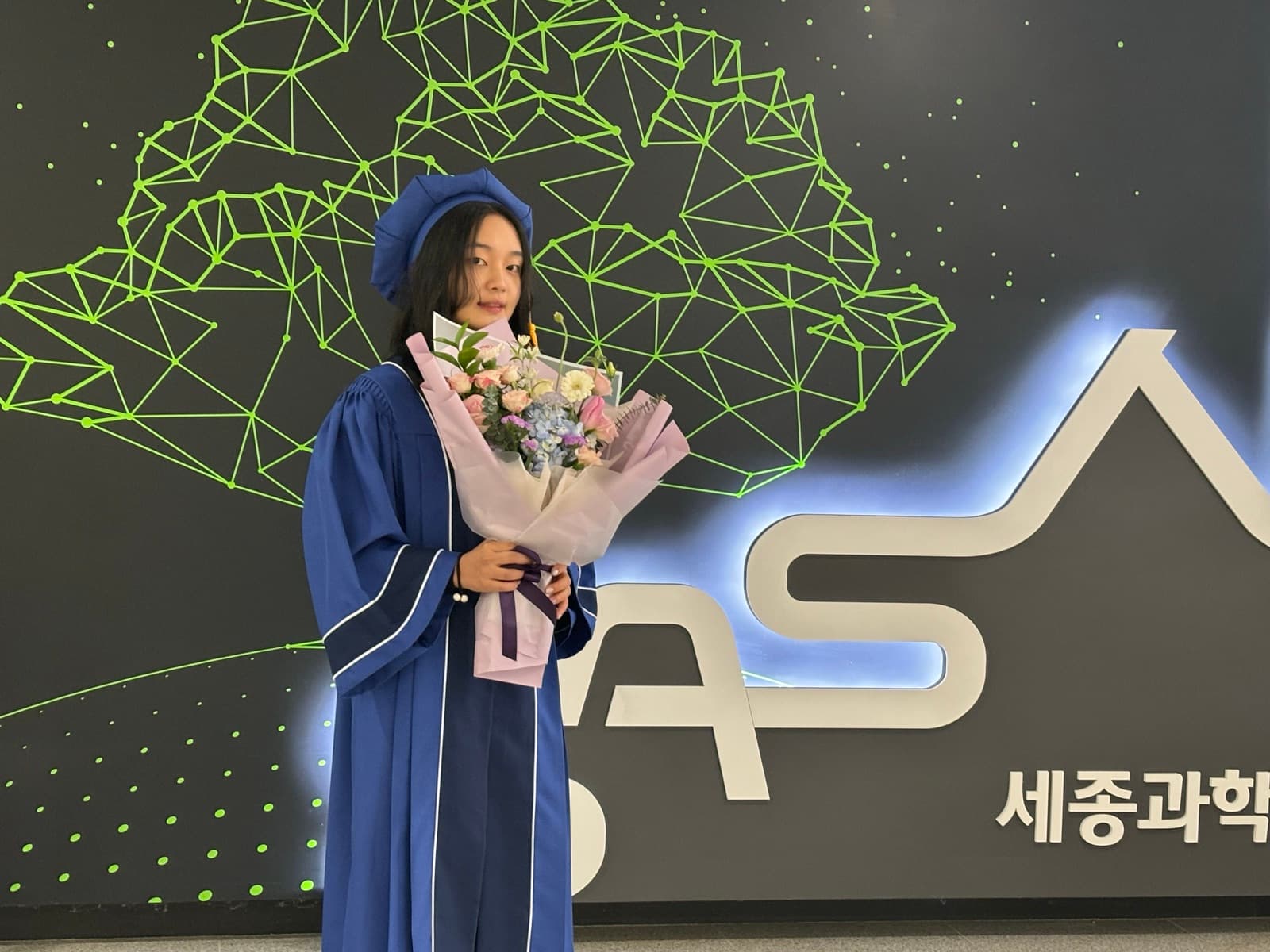 Nicole in a graduation cap and gown holding a bouquet of flowers in front of the Sejong Academy of Science and Arts (SASA) sign.