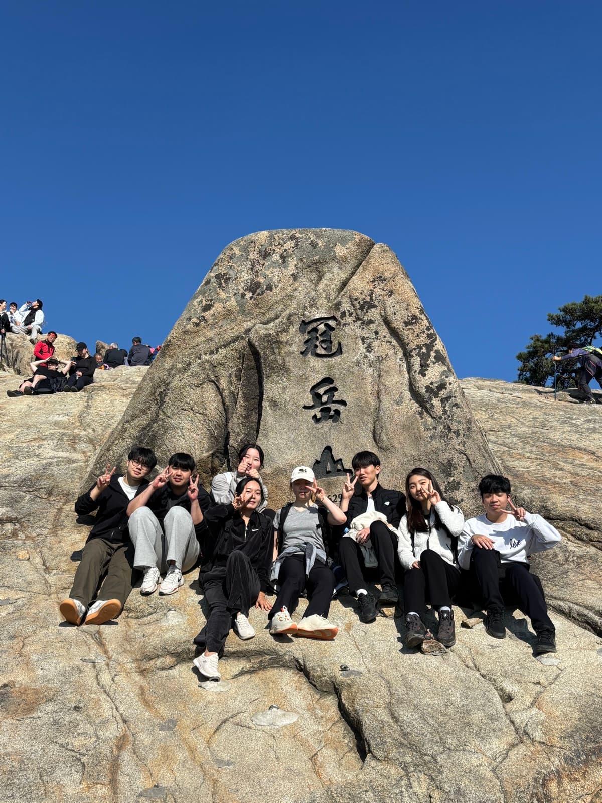 Nicole and friends sitting together in front of a large rock inscribed "관악산" (Gwanaksan) at the summit of the mountain next to SNU's campus.